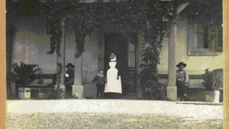 Photo de 1895 dévoilant  l’entrée de l’Hôpital Bon Vouloir, avec une Sœur donnant la main à un enfant  et deux hommes assis sur des banc accolés au mur. 