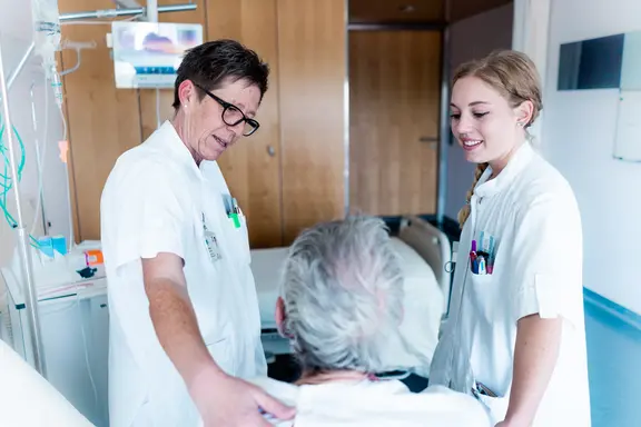 Photo d'une chambre d'hôpital. Deux infirmières discutent avec un patient vu de dos.